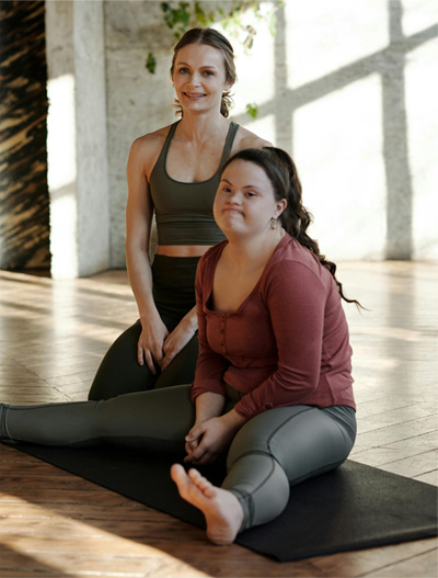 Image of two women, sitting on yoga mats and smiling at the camera